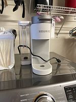 Front view of Keurig coffee maker showing the brand logo, white base, silver top, transparent water reservoir on the left, and power cord coiled in front.