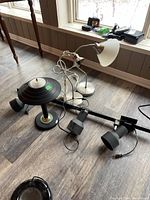 Photo showing all lamps and the track lighting fixture gathered on the floor near a window. The lamps include two white desk lamps, one black table lamp with a circular layered shade with gold accent, and a black track lighting bar with spotlights.