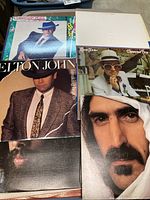 Stack of LP covers including Elton John in suit and hat alongside a white-label LP and a Frank Zappa close-up face