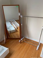 Full view of wood-framed floor mirror next to silver metal clothes rack against a wall on wooden floor