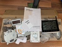 Photo showing multiple unassembled parts of the Oregon Scientific weather centre including display console, sensors, and manual inside a cardboard box.