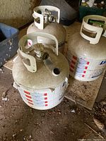 Three used Levaco propane tanks with valves and protective collars, placed on a wooden surface in a garage environment with visible dirt and debris