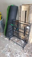 Photo showing three tiered black metal rack and rolled artificial grass carpet leaning against wall with other items in garage space.