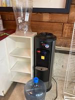 Photo showing the Vitapur black hot/cold water dispenser next to the white shelving unit and the clear plastic water container on top of the unit, with an empty large blue water jug in front.