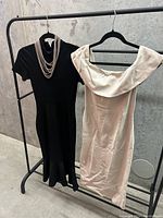 Two dresses hanging on black clothing rack against a concrete wall. Black short sleeve dress with layered silver necklace and off-white off shoulder dress with ruffled neckline.