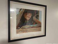 Framed print featuring a stylized portrait of a person resting their arms and head on a surface, shown hanging on a wall.