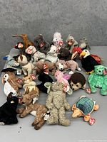 Photo showing 23 early Beanie Babies arranged on a gray surface, each with a Ty tag on the ear, featuring various animals and birds in different colors and textures.