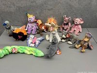 Photo showing thirteen assorted early TY Beanie Babies plush toys arranged on a gray surface, all with original TY tags.