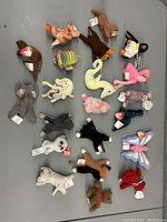 Top-down photo of 19 assorted early Beanie Babies plush toys laid out evenly on a gray surface showing detailed animal figures with original TY tags visible.