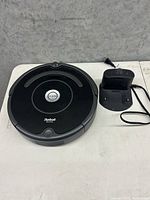 Top down photo of the black circular iRobot Roomba vacuum cleaner next to its compact charging station with attached power cord on a white surface.