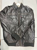 Front view of brown leather jacket showing zippered chest pockets, flap pockets, epaulets, and ribbed cuffs and waistband.