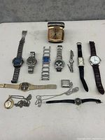 Photo showing a variety of wristwatches, two pocket watches with chains, a large travel alarm clock and one loose watchband displayed on a white surface.