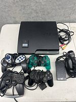 Top and angled view of Sony PS3 slim console with four wired controllers, power adapter, AV cables, and memory card on white surface.