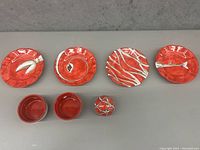 Top view of four hand painted red and white plates with two small red bowls and a decorative ceramic fruit piece, showing assorted designs including arrow, leaf, and wavy stripes.