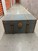 Full view of grey velvet or felt style chest with brown leather straps and decorative front clasp.