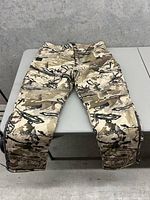 Front view of camouflage hunting pants laid flat on table showing knee stitching and leg zippers.