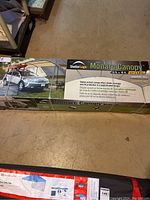 Top view of large sealed box for ShelterLogic Monarc Canopy showing model and specifications, including a front image display of the completed canopy covering a car.