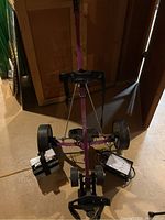 Full view of the purple battery powered golf cart showing two large rear tires, front wheels, footrest with securing strap, battery and charger placed on the floor nearby.