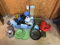 Assorted camping cookware including frying pan, plates, metal bowls, cups, and a basket with mugs and bottles