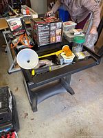 Black and silver small computer desk with assorted tools and hardware placed on top in a garage setting under natural lighting. View shows multiple items such as tools, funnel, containers, measuring tape.