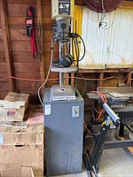 Full view of Rockwell Beaver drill press unit in garage setting showing gray metal base and drill head, power cable, and nearby boxes.
