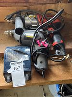 Photo showing a black and gray Black and Decker hammer drill with cord and plug, along with two other drills and power cords tangled together, plus a blue plastic case for drill bits labeled with Maxsold auction tag 167P.