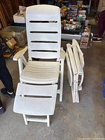 Two white plastic folding reclining chairs with folding footrests and three boxes of LED multi-color Christmas lights in a garage setting with other boxes and storage items.