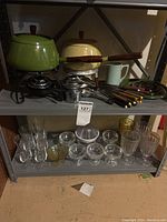 Overview of two fondue pots on upper shelf, assorted metal fondue forks, small green pitcher, and multiple small glasses on lower shelf.