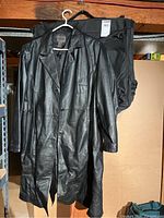 Black long women's leather coat hanging on a hanger with black garment travel bag behind it