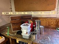 Overview of lot on table showing tool chest, plastic bins, metal cans, and large wooden level observed as primary items.