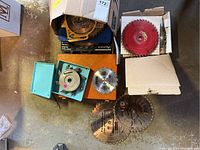 Various saw blades and discs arranged on garage floor, including boxed blades, red coated blade, and smaller blade in blue cases.
