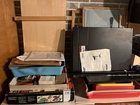 Overview of multiple stacked items including black panels, cardboard boxes, trays with papers and miscellaneous small items