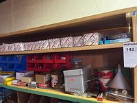 Shelf with white labeled Westcott Construction Hardware boxes, red and blue plastic bins filled with screws, nails, nuts, washers, bolts, metal brackets, and containers including a coffee tin and gray metal pocket office box.