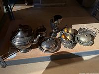 Photo of silver serving set including teapot, creamer, sugar bowl, covered dish, goblets, and handled cups on shelf