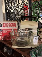 Vintage Sunbeam Mixmaster stand mixer with glass bowl and boxed slicer/shredder attachment placed on table with decorative metal background.