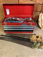 Front view of the red Husky metal tool box open with tools visible inside and three large hand saws in front.