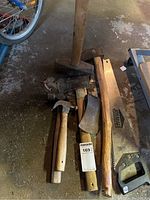 Photo showing assortment of tools including sledge hammer, axes, small hammers, and hand saw on garage floor.