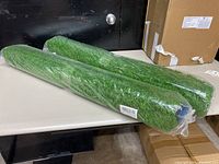 Two rolled green artificial turf rugs wrapped in clear plastic on a table.