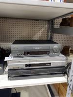 Three DVD VHS combo players stacked on a shelf, showing front panel details including buttons, input ports, and brand logos.