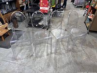Four clear lucite chairs arranged side by side showing full view. Chairs have oval backs, square seats, and straight legs. Floor surface and surrounding objects visible.