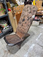 Angled side view of wooden chair showing seat and carved tall backrest with detailed wildlife and figural motifs.