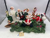 Wide angle photo showing full grouping including five Santa dolls in different outfits, reindeer, elfish figure in boat, and Christmas greenery base.