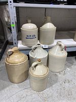 Six antique stoneware crocks and jugs on a concrete floor, showcasing different shapes, sizes, and glazes