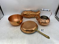 Four copper kitchen items displayed on a white surface: mixing bowl, frying pan, fish cake mold, and teakettle with porcelain handle.