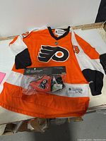 Complete view of the orange and white hockey jersey with Flyers logo and new pack of Flyers premium face masks on top.