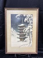 Full view of framed artwork showing a snow covered multi-level Japanese pagoda temple with trees and snowfall under a dark sky, matted and framed in brown ornamental frame.