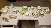 Wide overhead photo showing entire assortment of teacups, saucers, small bowls, and decorative china pieces arranged on a white table.