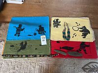 Four textile placemats displaying indigenous artwork in black on blue, yellow, green, and red backgrounds, with a beaded necklace placed on the yellow placemat.