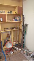 Wide view showing assortment of tools standing against wall including sledgehammer, axe, level, saws, and a green garden hoe still in packaging plus shelves with lubricant cans and work light on floor.