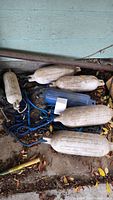 Photo showing a pile of white, blue, and some black boat bumpers on the ground with blue ropes attached, against a light green wall with fallen leaves on the floor.